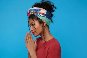 Positive young lovely dark haired curly woman with multi-colored headband smiling slightly while raising hands in praying gesture, standing over blue background photo