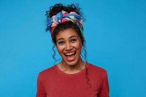 Overjoyed young lovely brunette curly female dressed in burgundy t-shirt and colored headband keeping her mouth wide opened while laughing cheerfully, isolated over blue background photo