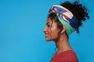 Side view of young attractive brunette woman with gathered curly hair looking cheerfully ahead with broad sincere smile while standing over blue background photo