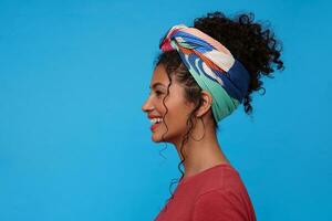 Side view of pretty young brunette curly woman with multi-colored headband looking cheerfully ahead with charming smile, isolated over blue background photo