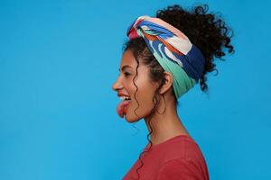 Funny shot of young joyful dark haired curly woman with festive makeup sticking cheerfully out her tongue while fooling, standing over blue background photo