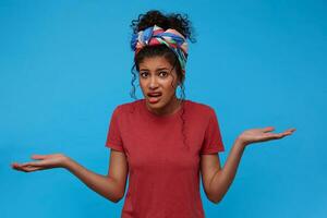 Confused young brown-eyed curly female with multi-colored headband keeping palms raised and grimacing perplexedly face while posing over blue background photo