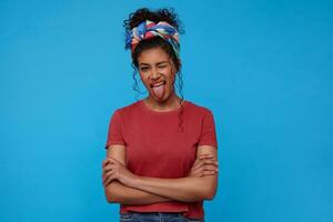 Joyful young lovely brunette curly female with festive makeup keeping one eye closed while sticking out her tongue, posing over blue background with crossed hands photo