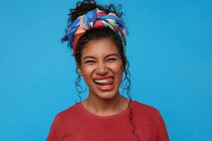 Portrait of young joyful dark haired lady with nose piercing squinting her eyes and showing cheerfully tongue while standing over blue background in colored clothes photo