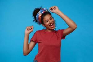 Joyful young attractive brunette curly woman with multi-colored headband singing and dancing cheerfully while standing over blue background with raised hands photo