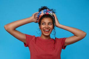 Joyful young attractive dark haired female with multi-colored headband looking happily aside with broad smile and keeping hands raised while posing over blue background photo