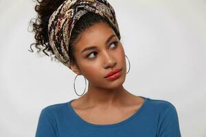 Indoor shot of young pretty dark haired curly lady tilting her head while looking wonderinly aside, dressed in blue blouse and colored headband while posing over white background photo