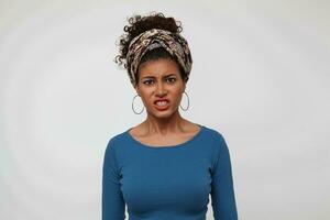 Indoor photo of young irritated dark haired curly lady with casual hairstyle grimacing her face while looking discontentedly at camera, standing over white background