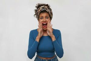 Agitated young dark haired curly woman with dark skin raising hands to her mouth while screaming excitedly with wide mouth opened, isolated over white background photo