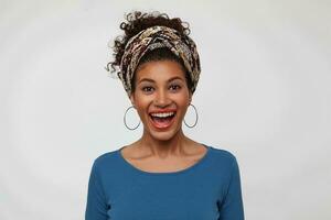 Studio photo of happy young dark haired curly trendy lady laughing cheerfully while rejoicing about something, isolated over white background with hands down