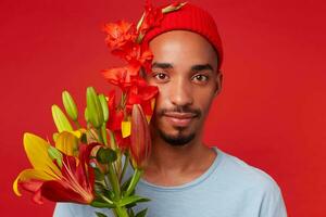 cerca arriba de joven atractivo chico en rojo sombrero y azul camiseta, sostiene un ramo de flores en su manos, mira a el cámara con calmante expresión y sonriente, soportes terminado rojo fondo foto