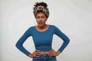 Bewildered young brown-eyed brunette dark skinned woman keeping hands on waist while looking scowlingly at camera, standing against white background photo