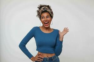 Studio shot of young joyful dark haired curly lady dressed in trendy colored clothes raising hand while looking cheerfully aside with wide mouth opened, isolated over white background photo