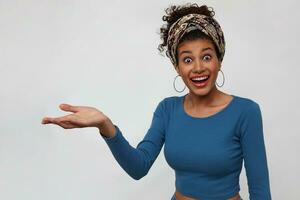 Studio shot of surprised young pretty dark haired curly lady rounding amazedly her brown eyes while showing aside with raised palm, isolated over white background photo