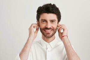 de cerca de contento hermoso joven empresario con cerda y inalámbrico auriculares usa camisa mira seguro, sonriente y escuchando a música aislado terminado blanco antecedentes foto