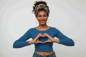 Happy young attractive brunette dark skinned woman showing love sign with raised hands and smiling cheerfully at camera while standing over white background photo