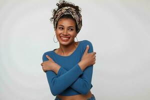 Cheerful young attractive brunette female with colored headband hugging herself with raised hands and smiling happily while standing over white background photo