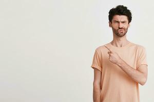 Portrait of unhappy displeased young man with bristle wears peach t shirt looks upset and points to the side with finger isolated over white background photo