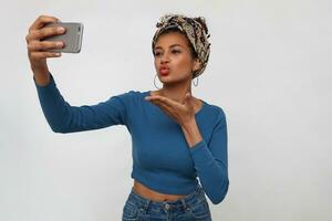 Attractive young brunette dark skinned female sending air kiss at camera while making photo of herself on mobile phone, standing over white background