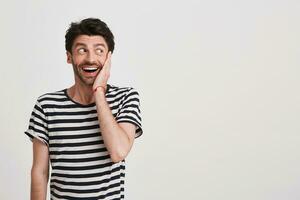 Closeup of surprised excited young man with bristle and hand on cheek wears striped t shirt feels happy and looks to the side isolated over white background photo