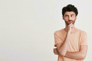 Portrait of serious thoughtful young man with bristle wears peach t shirt looks pensive and keeps hands folded isolated over white background photo