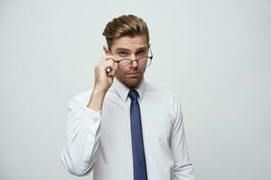 atractivo hermoso empresario con elegante Corte de pelo, pone lentes abajo a el propina de el nariz a ver algunos detalles, vestido en perfectamente planchado blanco camisa y azul corbata, encima blanco antecedentes foto