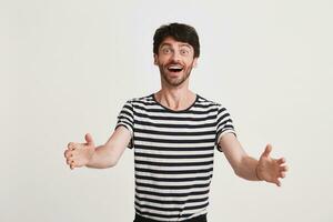 Portrait of cheerful amazed young man with bristle wears striped t shirt and opened mouth feels excited and ready to hug you isolated over white background photo