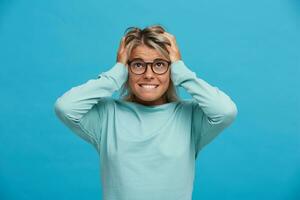 Shocked scared blonde girl, looks up frightened afraid of the imagine consequences of her actions, bites lower lip, hands holding head, over blue background photo