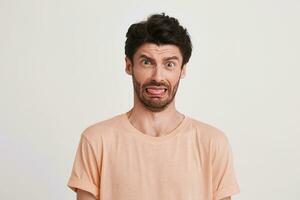 Closeup of funny dissatisfied young man with bristle wears peach t shirt looks displeased and shows tongue isolated over white background photo