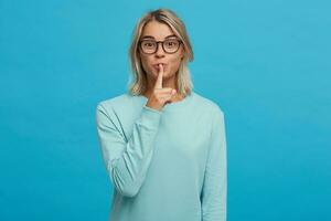 Emotional young girl in glasses calls for silence, keep a secret, do not tell anyone, keep quiet, do not make noise, keeps fore finger on lips, demonstrates silence gesture, on a blue background photo