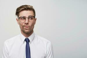 Young european man thinks, looks through glasses aside incredulously, with doubt, one corner of the lips pursed, white background, blank copy space for text or advertisement photo