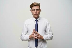 A young man stands with his fingers folded in front, looks through glasses, a little nerdy, tense, tries to make a choice or find a problem decision, on a white background photo