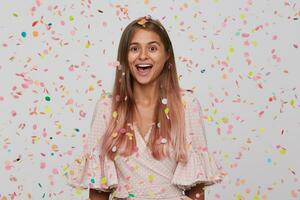 Portrait of cheerful beautiful young woman with long dyed pastel pink hair wears polka dot pink dress feels excited, having party and shouting isolated over white background with confetti photo