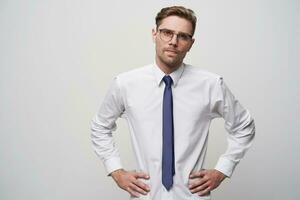 un joven hombre en un duro pose, mira mediante anteojos, manos en caderas, lo hace no Mira muy complacido, insatisfecho, pretende a cambio algo, a Rápido su opinión, en un blanco antecedentes foto