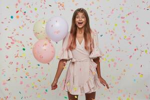 young woman with long dyed pastel pink hair and opened mouth wears polka dot pink dress celebrating birthday, holding colorful baloons in hand isolated over white background with confetti photo