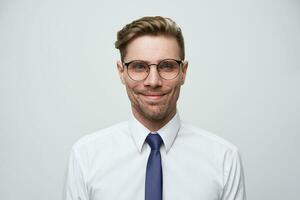 Calm interesting unshaven guy in white shirt and blue tie with smiling face expression, looking at camera while standing against white background. Man takes photo for passport at local studio