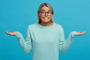 Confused young girl shrugging shoulders, having guilty look, feeling sorry for doing something wrong and making terrible mistake, dressed in light blue long sleeve t-shirt, over blue background photo