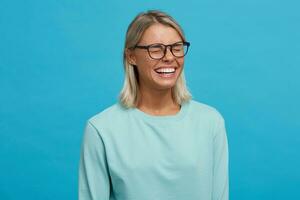 Indoor shoot of funny cheerful glad blonde young girl in glasses, widely smiles, closes eyes while laughing, standing sideways, dressed in light blue long sleeve t-shirt,on a blue background photo