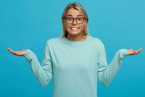 Guilty and confused young girl shrugging shoulders, making helpless gesture with hands, having Oops expression on her face. Feeling sorry for doing something wrong,over blue background photo