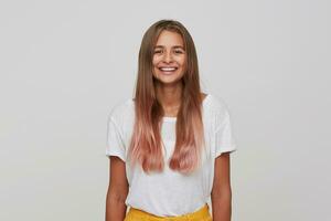 Portrait of cheerful beautiful young woman with long dyed pastel pink hair feels happy and smiling isolated over white background Looks directly in camera photo