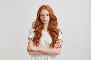 Portrait of serious beautiful redhead young woman with long wavy hair and freckles wears t shirt keeps arms crossed and feels confident isolated over white background photo