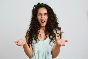 Portrait of angry mad young woman with long dark curly hair and opened mouth shouting and holding copy space on both palms isolated over white background Feels irritated photo