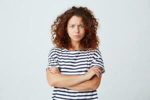 Closeup of serious worried young woman with curly hair wears striped t shirt feels embarrassed and stands with arms crossed isolated over white background photo