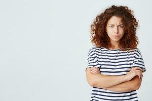 Closeup of angry stressed curly young woman wears striped t shirt have problems and stands with arms crossed isolated over white background photo