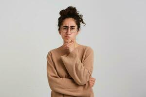 Thoughtful concentrated young woman with bun of dark curly hair wears beige pullover and glasses keeps hands folded and thinking about her future job isolated over white background photo