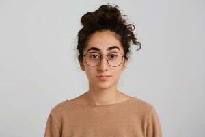 Closeup of serious pretty young woman with bun of dark curly hair wears beige sweatshirt and glasses isolated over white background Looks directly in camera photo