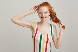 Closeup of cheerful lovely redhead young woman with freckles and ponytail wears striped top, touching her hair and posing isolated over white background Feels happy photo