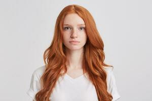Closeup of serious beautiful redhead young woman with long wavy hair and freckles wears t shirt feels confident and looks directly in camera isolated over white background photo