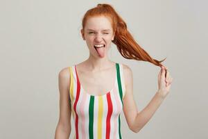 Closeup of funny cute young woman with red hair and freckles in striped top winking, showing tongue and having fun isolated over white background photo