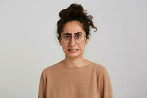 Portrait of embarrassed worried young woman with bun of dark curly hair wears beige sweatshirt and glasses feels confused and concerned isolated over white background photo